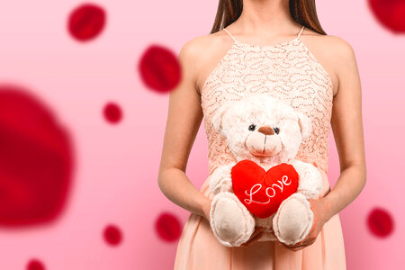 A young girl in a pink dress holding a teddy bear with a heart of love. Pink background. Blured rose flowers. photo