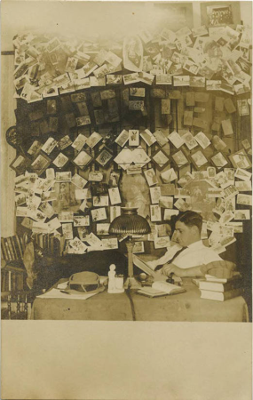 Man sitting at a desk, reading, and a lot of postcards hanging in the background (NBY 422994)