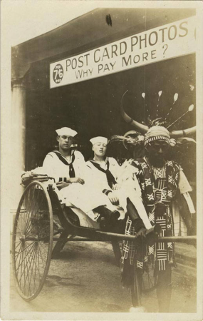 Two sailors, sitting in a man-pushed cart, posing in front of a Post Card Photo shop (NBY 422542)