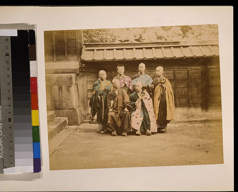 Six men, possibly monks, posed for group portrait, four standing and two sitting in front, five have their heads shaved, wearing geta, some are holding items, with building in background LCCN2009632883, 1877