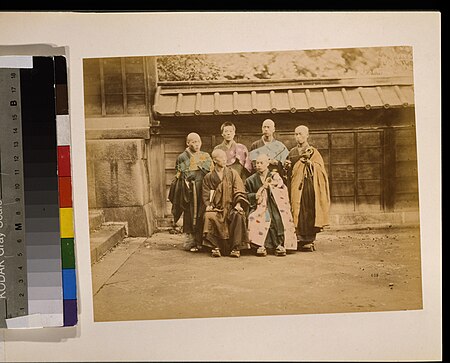Six men, possibly monks, posed for group portrait, four standing and two sitting in front, five have their heads shaved, wearing geta, some are holding items, with building in background LCCN2009632883, 1877