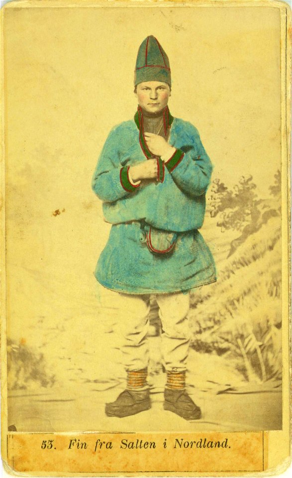 53. Mannsdrakt, samedrakt, Salten. Portrett av mann stående i fotoatelier foran malt naturbakgrunn - Norsk folkemuseum - NF.08676-004 (1)