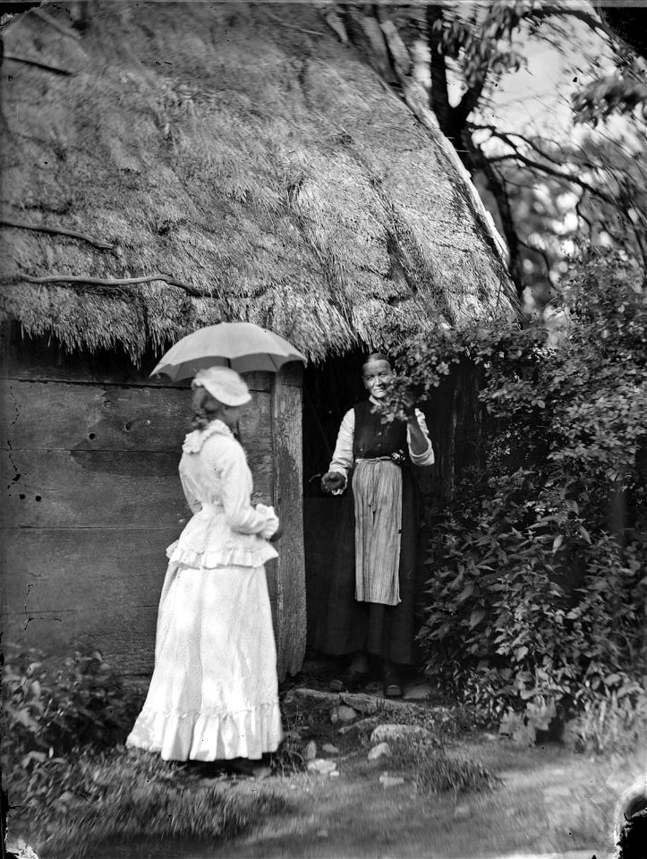 En äldre kvinna står i dörröppningen till en stuga. Halland - Nordiska Museet - NMA.0052613
