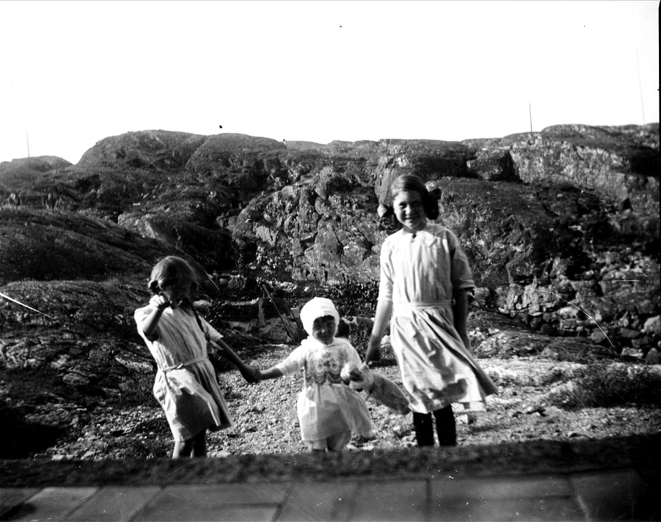 Tre flickor iklädda klänningar håller varandra i handen på strand. Troligen i Fiskebäckskil, Bohuslän - Nordiska Museet - NMA.0054136