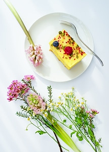 Slice of Cheesecake Decorated with Fruits Surrounded by Flowers on White Table