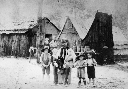 Father and children outside their home. Lady and gentleman in background, [n.d.] photo