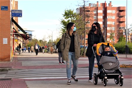 Plano general de dos mujeres con mascarilla paseando por la acera con un carrito de bebé con un niño dentro
