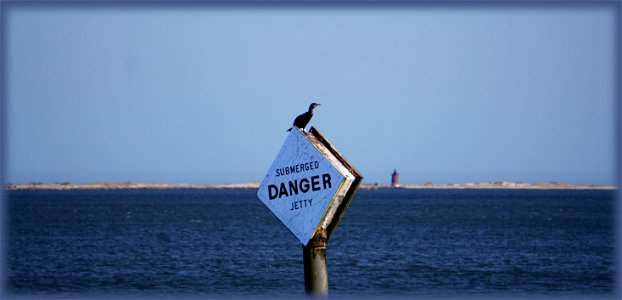 Warning sign for jetty near Cape Henlopen. A warning sign, breakwater, and East End lighthouse are in the photograph.