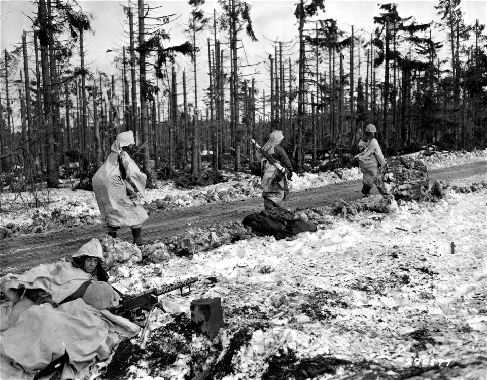 SC 270597 - Camouflaged infantrymen of the 9th Infantry, 2nd Division, pass an American light machine gun position and a dead German as they advance along a road in the Monschau Forest, Germany. 3 February, 1945.