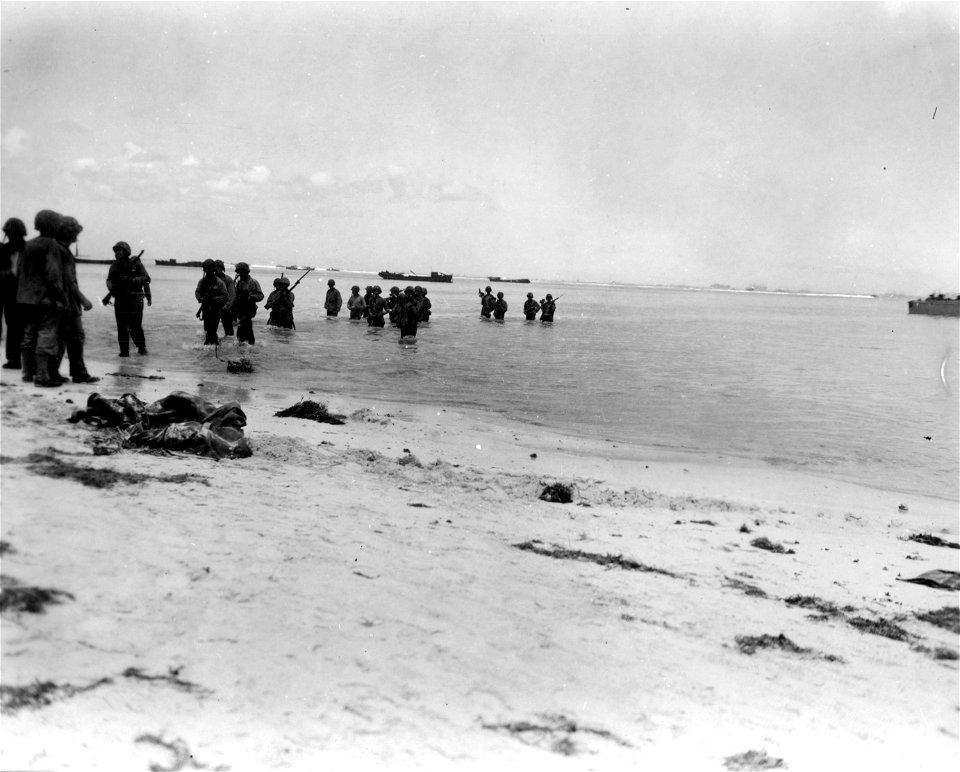 American troops, their landing boat 50 yards from the water's edge, wade ashore, rifles and other weapons carried overhead, joining the assault as reinforcements on beaches of Saipan, 1st day of the attack.