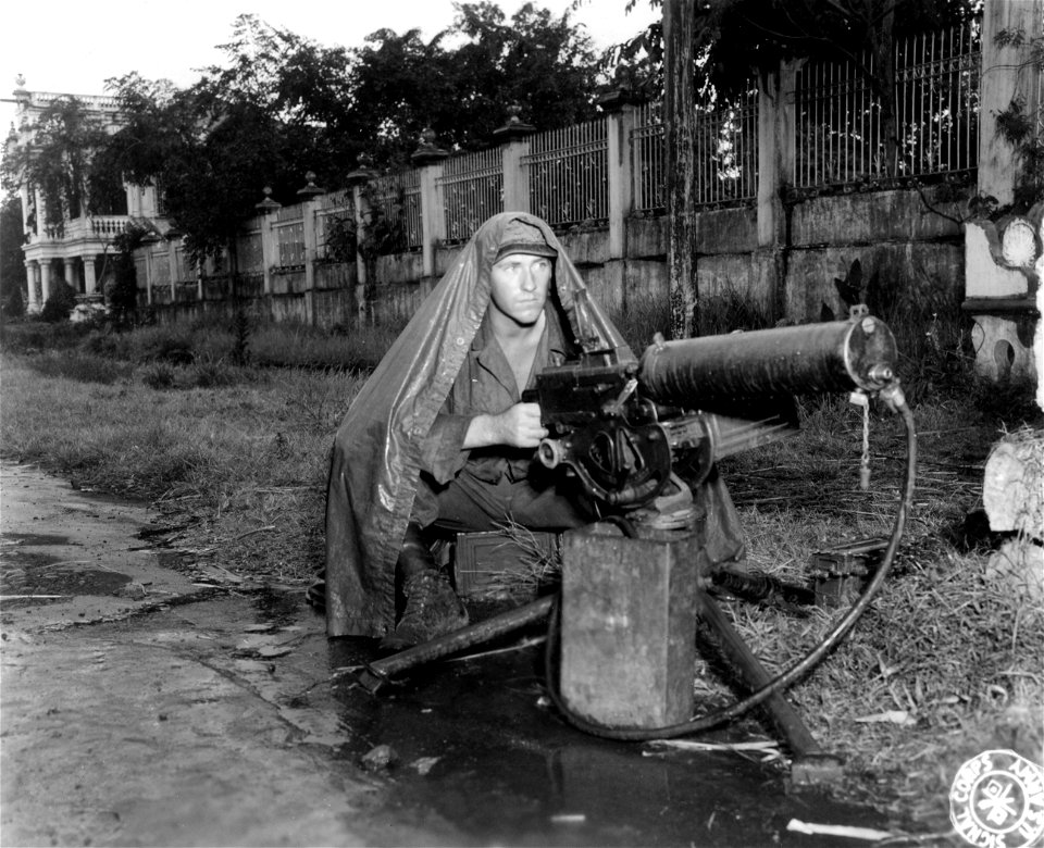 SC 364514 - Pfc. Thomas Powell, North Platte, Nebr., 5th Cav., guards the main street of San Pablo, Luzon, P.I, with his machine gun, and covers himself completely with his poncho in the tropical rain.