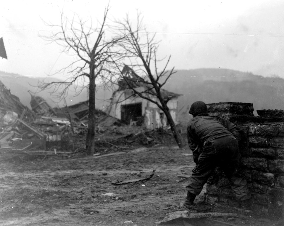 SC 270605 - An American infantry soldier of the 2nd Infantry Division, 1st U.S. Army, crouches behind cover of a wall as he watches the taking of a German pillbox on high ground overlooking the town of Gemund, Germany. 4 March, 1945.