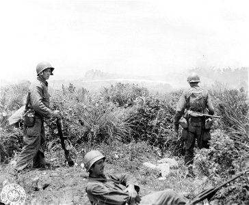 SC 270802 - Three riflemen of the 32nd Regiment, 7th Infantry Division, remain at a safe distance while a flame-throwing tank in the background sprays a Jap emplacement. 19 June, 1945. photo