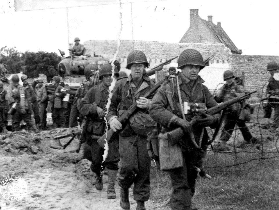 SC 270658 - American troops move into French village supported by a tank in the background. Ever increasing numbers of well trained, expertly equipped men are pouring in to oppose Hitler's forces on the Continent. 9 June, 1944.