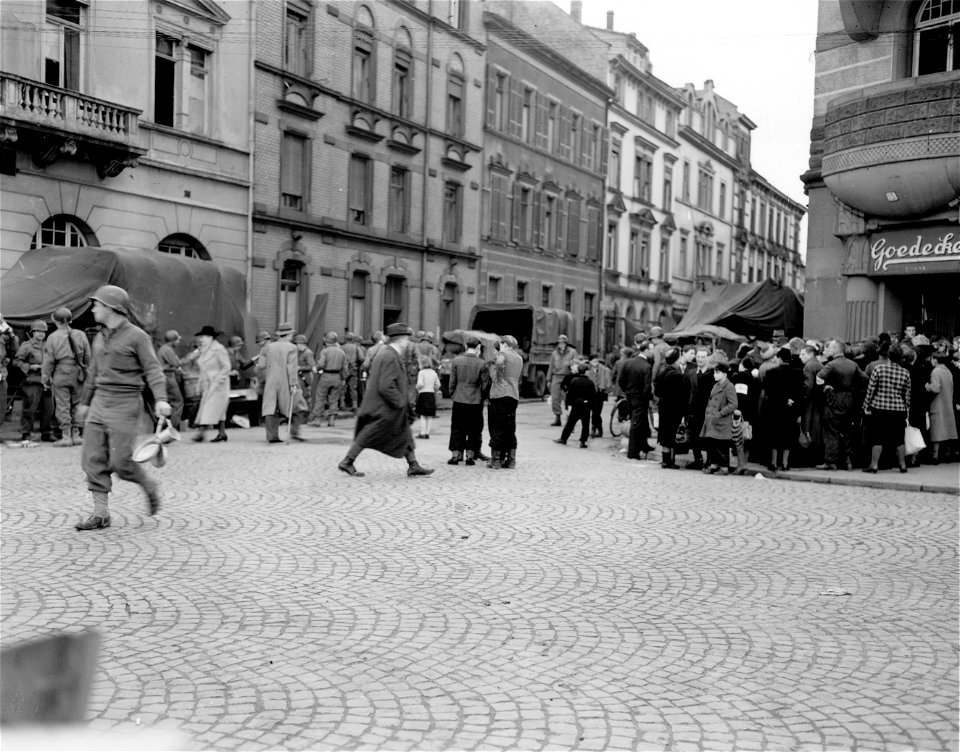 SC 335289 - German civilians of Heidelberg stand waiting for their ration of bread while soldiers across the street get their fill of chow.