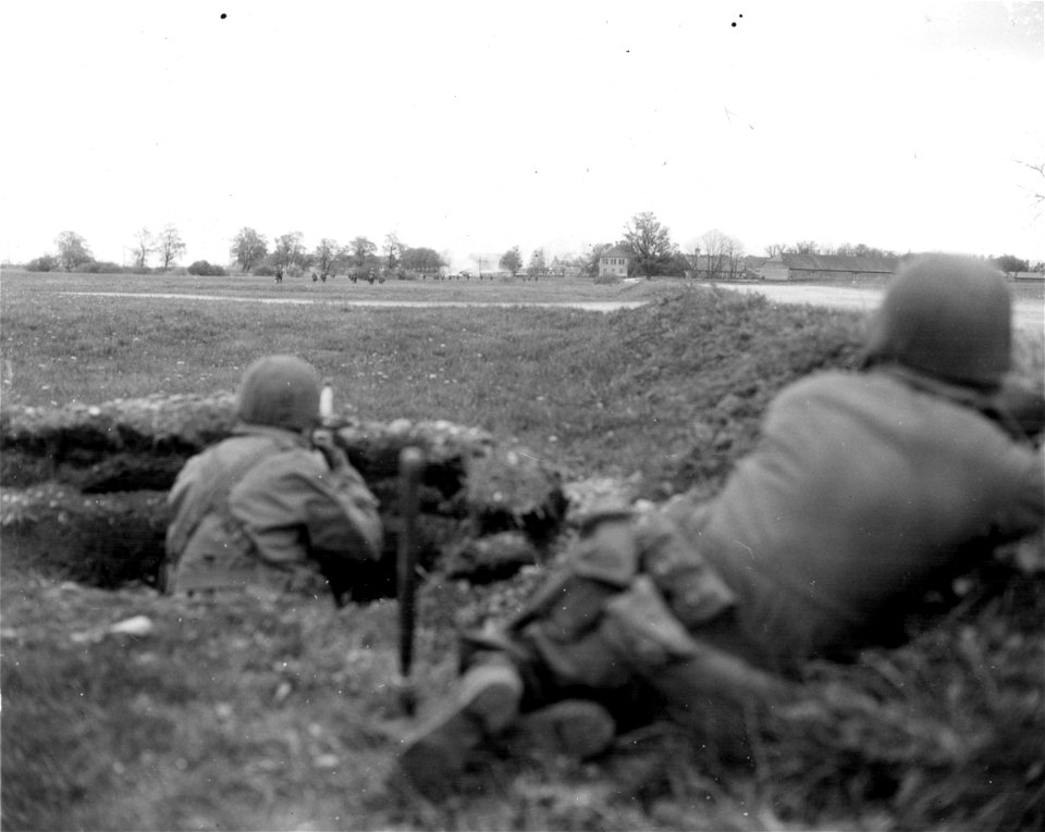 SC 270648 - Two infantrymen in foreground in foxhole and lying along bank, support the forward elements in the distance as they move into small burning town, a strongpoint on the march to Munich.