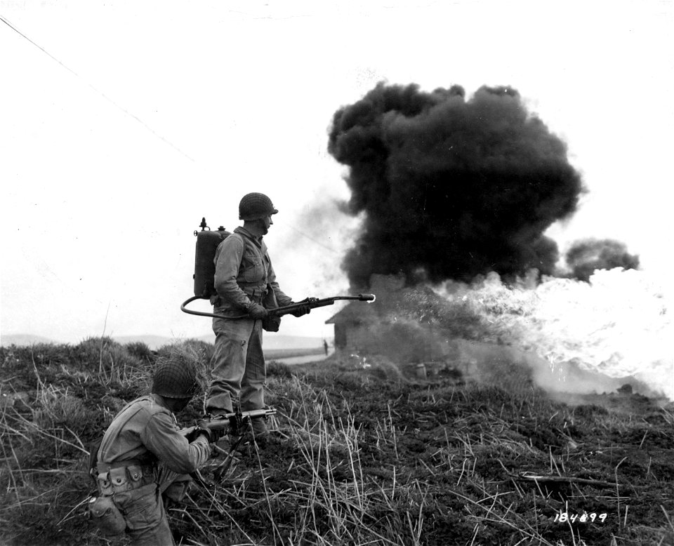 SC 184599 - Pfc. Elmar Kuell(?), 29, Spring Valley, Wisc., demonstrates his flame-throwing apparatus while Pfc. Chas(?) Paterson, Clovis, New Mexico, guards for him.