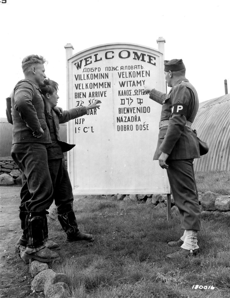 SC 180016 - Sgt. Blackmon explains the welcome sign in fifteen languages near the entrance to a camp in Iceland. Gefr. Lehn Obergefr. Klinkmann view the sign with a show of interest.