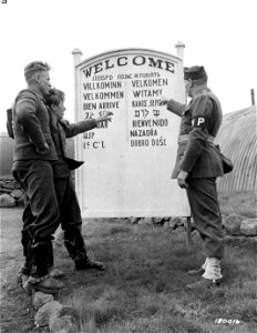 SC 180016 - Sgt. Blackmon explains the welcome sign in fifteen languages near the entrance to a camp in Iceland. Gefr. Lehn Obergefr. Klinkmann view the sign with a show of interest. photo