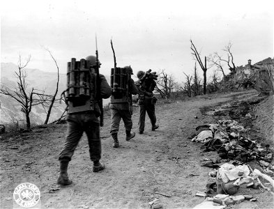 SC 374760 - Past some strewn about German equipment, and a dead German soldier that someone thoughtfully covered up, go three men carrying howitzer ammunition to the top of the mountain. Mt. Belvedere area, Italy, 21 February, 1945.
