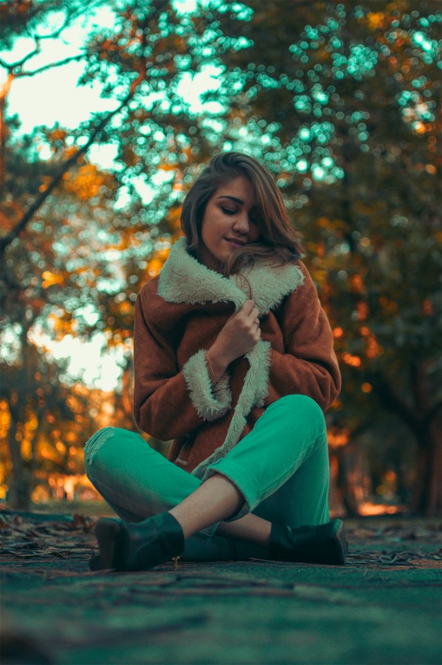 Woman Wearing Brown-and-white Coat And Teal Denim Jeans Sitting On Road Surface