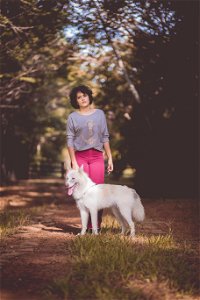 Woman In Purple Long Sleeve Shirt And Pink Jeans Standing Next To White German Spitz With Background Of Green Trees During Day photo