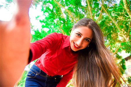 Smiling Woman In Red Shirt And Blue Jeans Taking Selfie Under Green Leaved Tree photo