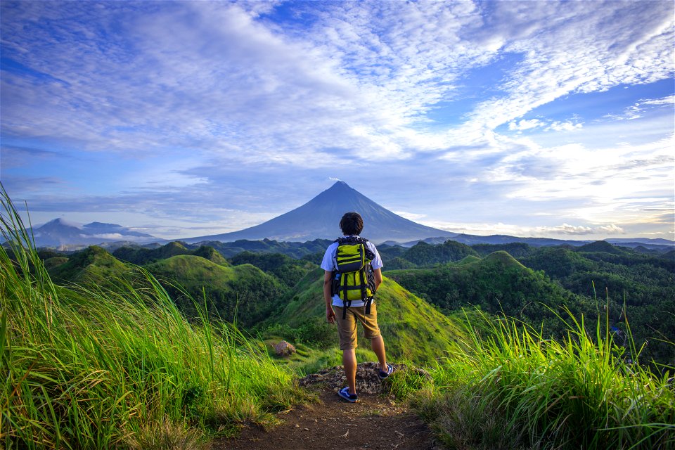 Man Wearing White Shirt Brown Shorts And Green Backpack Standing On Hill