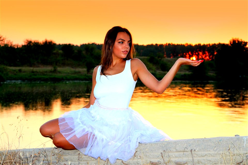 Woman In White Square Neckline Sleeveless Dress Sitting On Beige Wall Beside Body Of Water During Golden Time