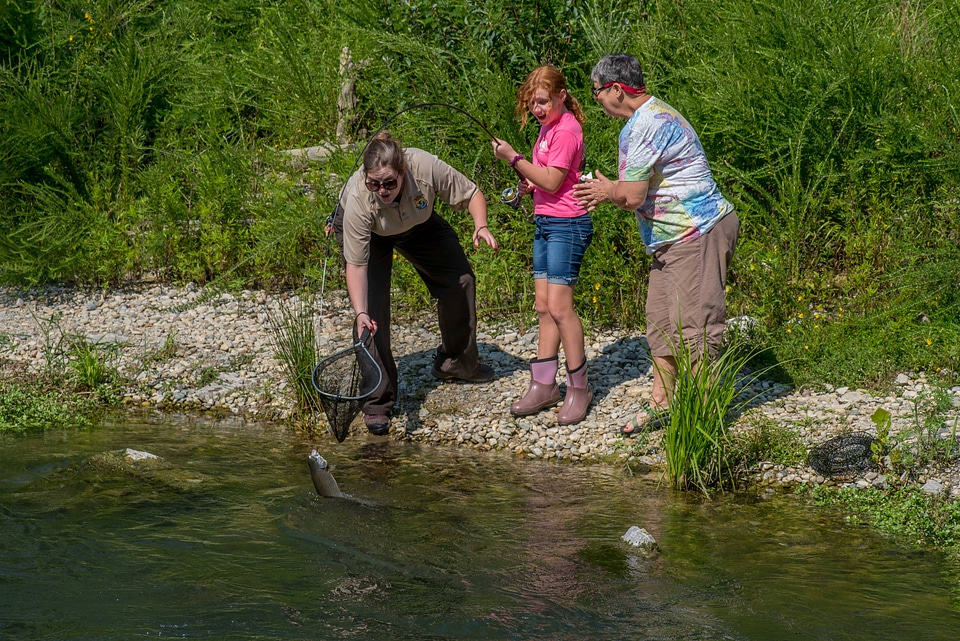Older woman fishing with young redheaded girl, FWS employee netting trout-1