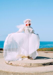 Woman in White Lace Wedding Dress Standing on Gray Pavement Near Body of Water during Daytime photo