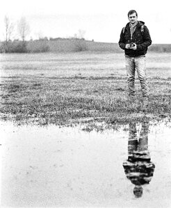 Man in Jacket Standing Near Body of Water Holding Camera in Grayscale Photography photo