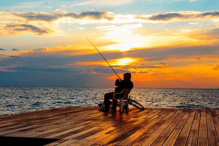 Silhouette of Person Sitting on Chair Holding Fishing Rod Near Body of Water during Golden Hour photo