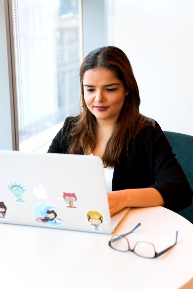 Woman Wearing Black Suit Jacket Using White Macbook With Stickers On White Wooden Table
