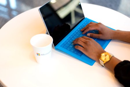 Person Wearing Round Gold-colored Watch Using Black Tablet Computer With Blue Detachable Keyboard On Round White Wooden Table photo