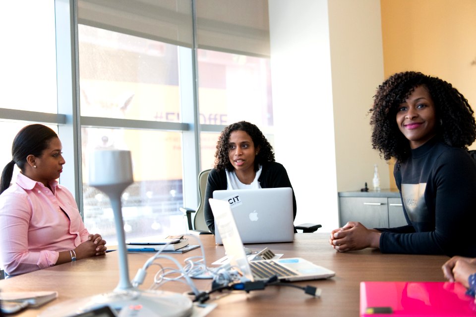 Three Woman Sitting Beside Brown Wooden Conference Table With Silver Apple Macbook