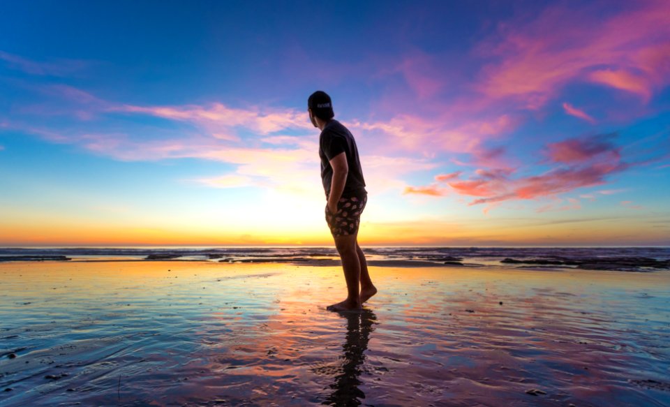 Man In Black T-shirt Wearing Cap Near Body Of Water During Sunset