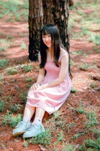 Girls Pink Sleeveless Dress Sits Beside Black Tree Trunk At Daytime