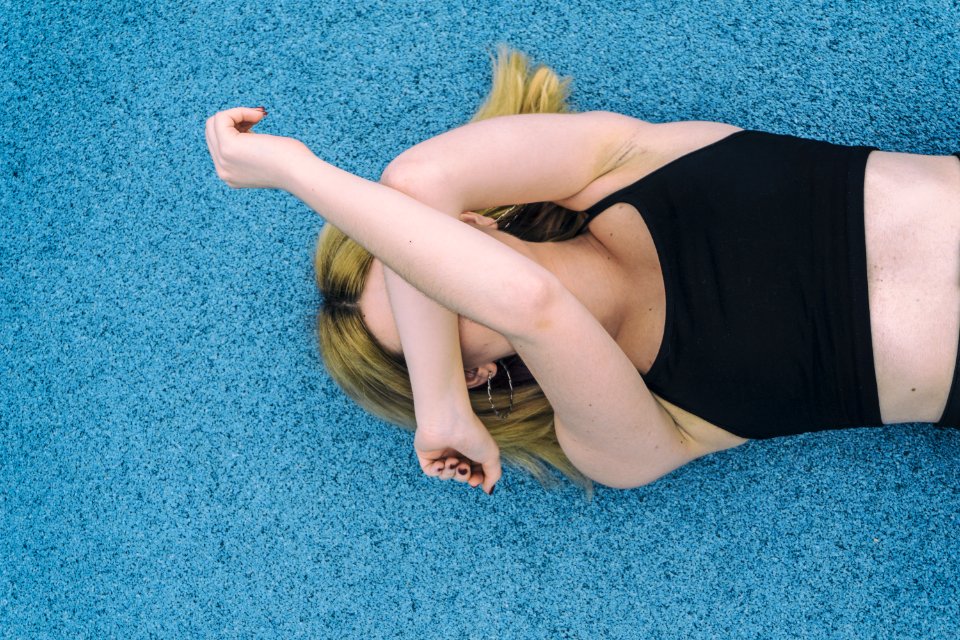 Woman Wearing Black Sports Brassiered Lying On Teal Surface While Covering Her Face With Her Arms
