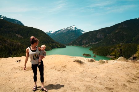 Woman Wearing Grey Tank Top Carrying Baby In Distant Of Lake Between Mountains photo