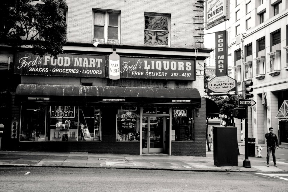 Greyscale Photo Of Man Standing On Road Near Freds Liqours Building