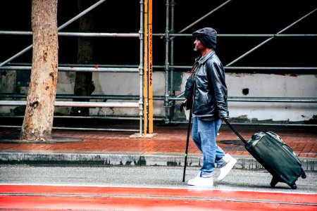 Walking Man In Black Leather Jacket And Blue Denim Pants Holding Luggage Bag photo
