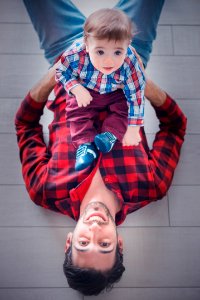 Lying Man Wearing Red And Black Gingham Long-sleeved Shirt Holding Toddler Sitting On His Stomach