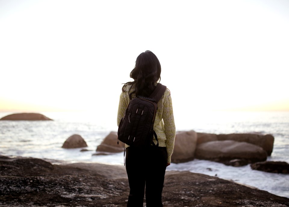 Woman Wearing White Long-sleeved Top And Black Pants Carrying Black Backpack While Standing On Shore During Adytime