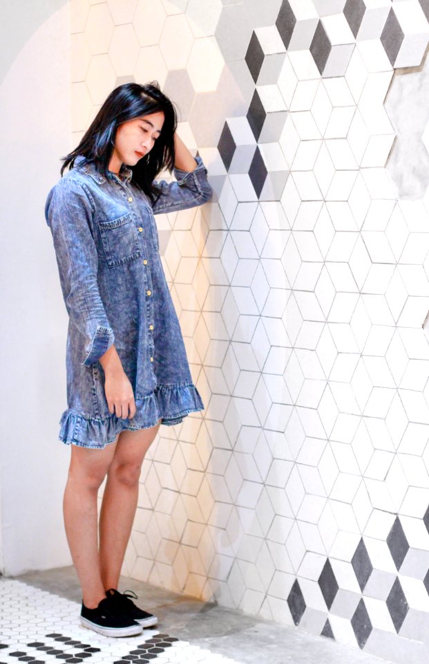 Woman Wearing Blue Denim Button-up Long-sleeved Dress Holding Her Head With Her Left Hand Standing Near White And Gray Concrete Wa