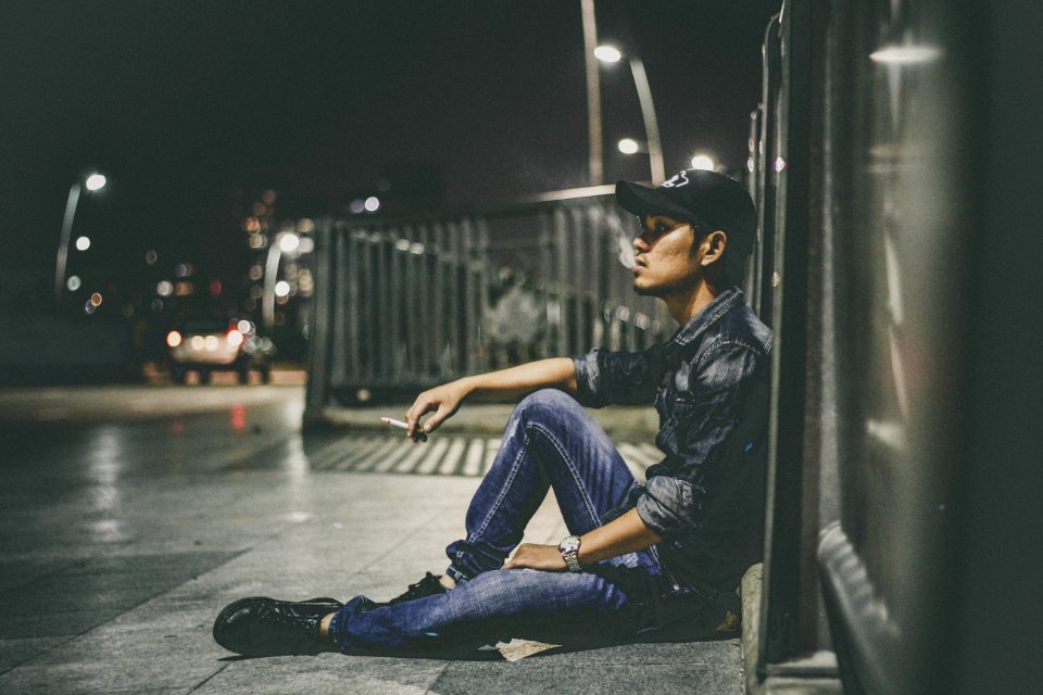 Shallow Focus Photography Of Man Sitting Beside Railing Holding Cigarette Stick