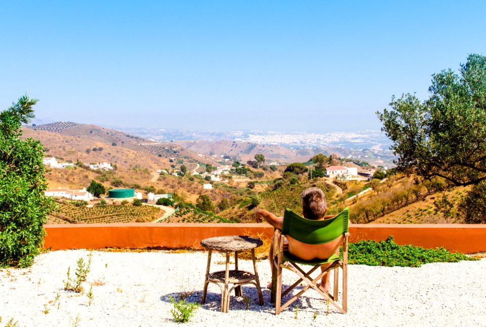Man Sitting On Green Chair Near Trees And Mountain Under Blue Sky At Daytime
