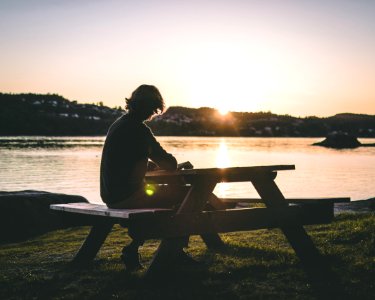 Silhouette Of Person In Black Top Sitting On Picnic Bench Near Body Of Water During Sunset photo