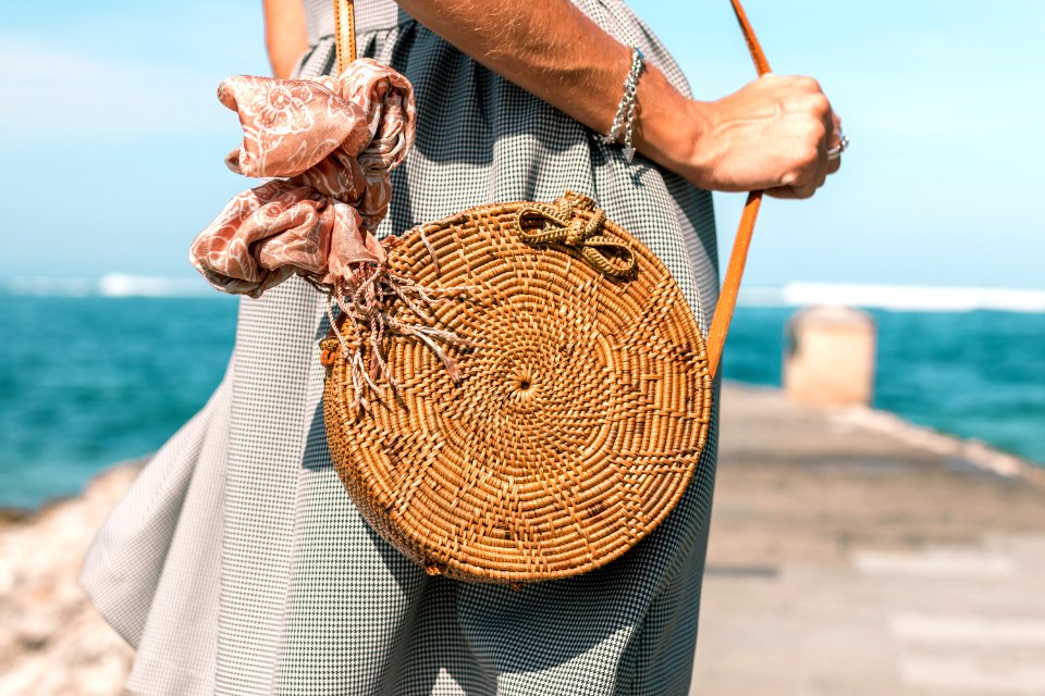 Woman Wearing Grey Skirt And Round Brown Rattan Crossbody Bag On Wooden Dock Near Body Of Water