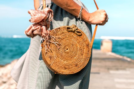 Woman Wearing Grey Skirt And Round Brown Rattan Crossbody Bag On Wooden Dock Near Body Of Water